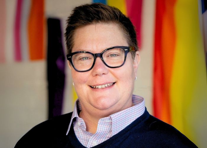 Tracy Hall is the new Executive Director of Affirmations LGBTQ+ Center in Ferndale MI. Tracy is a white nonbinary person with short hair, glasses, and wearing a collard shirt with a blue sweater. She is pictured in front of a variety of gay pride flags.